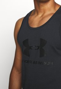 Zbliżenie na osobę w ciemnej bezrękawnikowej koszulce sportowej Under Armour z logo i nazwą marki na klatce piersiowej.