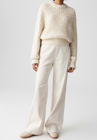 Personne portant un pull en grosse maille crème, un pantalon large en velours blanc et des chaussures décontractées beiges, debout devant un fond uni.