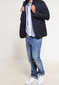 Marineblauer gesteppter Blazer mit einem kragenlosen Hemd darunter, hellblau mit weißen Punkten, kombiniert mit ausgewaschenen blauen Jeans und weißen Sneakern.