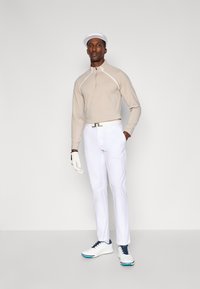 Beżowy pulower z długim rękawem i półzamkiem, białe akcenty; białe spodnie garniturowe; białe buty z granatowymi sznurówkami; biała czapka; rękawica golfowa na lewej ręce.