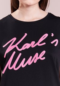 Černé tričko s krátkým rukávem, na přední straně s výrazným růžovým kurzívním textem „Karl's Muse“. Hladká látka s klasickým designem kulatého výstřihu.