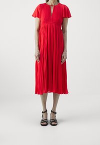 Abito rosso plissè con maniche corte a sbuffo e scollatura a V profonda; abbinato a sandali neri con lacci. Tessuto morbido con una silhouette fluida.