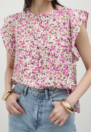 Frau trägt eine pink-beige geblümte Kurzarmbluse, die in hoch taillierte blaue Jeans gesteckt ist, mit goldenen Armreifen an beiden Handgelenken.