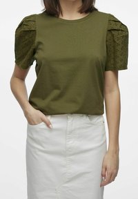 Olivgrön t-shirt med korta ärmar i öglemmönster, ihop med en vit denimkjol. Toppens halsringning är rund och har en avslappnad passform.