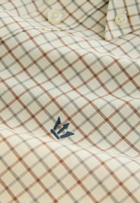 Beige ternetstof med brune og grå linjer; har et blåt broderet logo i nederste hjørne, der viser et afslappet skjortedesign.