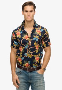Camicia a maniche corte blu navy con fiori grandi rossi e blu, foglie verdi e colletto a punta, abbinata a denim chiaro e una cintura marrone.