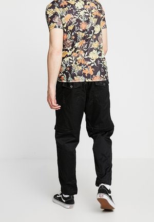 Persona che indossa una camicia a maniche corte con motivo floreale, pantaloni cargo neri e scarpe da ginnastica bianche e nere, che cammina su uno sfondo semplice.