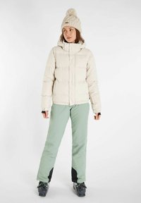 Veste beige rembourrée avec capuche, associée à un pantalon de ski vert clair. Bonnet en tricot crème avec un pompon. Bottes de ski noires.