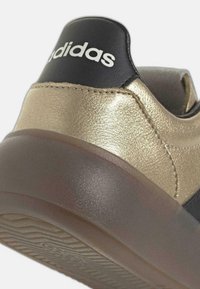 Primer plano del talón de una zapatilla metálica dorada con cuello negro y suela translúcida marrón, que presenta el logo blanco de Adidas en la pestaña trasera.