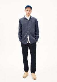 Marineblauwe overhemd met lange mouwen en knopen over een wit shirt, gecombineerd met zwarte broek en beige moccasin-achtige schoenen. Een blauwe pet maakt de look compleet.