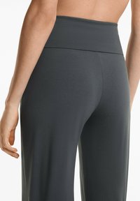 Nahaufnahme eines Rückens einer Person, die hochgeschnittene, dunkelgraue Leggings trägt, wobei ein Teil des Arms und des Oberkörpers vor einem schlichten Hintergrund zu sehen ist.