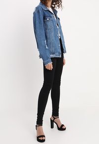 Vaate denim-takkia vaaleansinisellä värillä, johon on lisätty kulutustyylisiä yksityiskohtia. Takki on yhdistetty mustiin kapeisiin farkkuihin ja mustiin remmillä varustettuihin korkokenkiin.