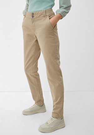 Beige recht gesneden broek met twee knopen, gedragen met witte sneakers en een lichtgroene top, handen in de zakken.