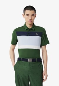 Grön pikétröja med marinblå, ljusblå och vita ränder. Bärs med gröna byxor med små logomönster och ett marinblått bälte.