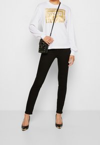 Bílá košile s dlouhým rukávem a zlatým logem Versace, kombinovaná s černými skinny džíny a černými vysokými podpatky. Součástí je také černá kabelka s pyramidovými nýty.