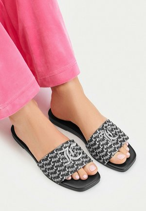 Piedi che indossano sandali slide neri con motivo a scrittura bianca e grande lettera decorativa T sulle fasce, abbinati a pantaloni rosa.