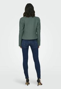 Veste verte texturée avec coupe ajustée, taille cintrée, manches longues et fente au dos, associée à un jean skinny bleu foncé et des talons noirs.