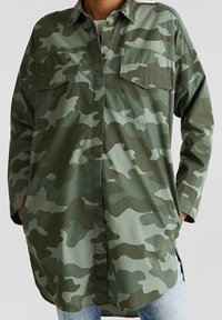 Chaqueta de camuflaje de manga larga y oversize con bolsillos en el pecho, llevada sobre una camisa blanca y jeans azul claro, con las manos en los bolsillos laterales.