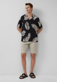 Chemise noire à manches courtes avec imprimé de feuilles de palmier blanches, short beige et sandales noires à boucles. Le modèle se tient avec les mains derrière le dos.