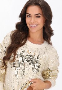 Krém színű pulóver, melyet arany és ezüst flitterek díszítenek, kerek nyakkivágással és puha textúrával rendelkezik, bordás mandzsettákkal és aljjal.