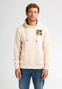 Sudadera de color crema con un bolsillo canguro, cordones ajustables y bordados geométricos multicolores en el pecho. Material suave de mezcla de algodón.