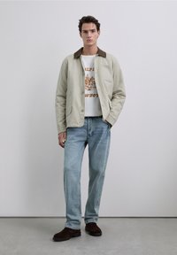 Beige jacket with brown collar, white graphic t-shirt featuring orange print, light blue denim jeans, and brown suede boots. Simple design.