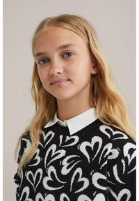 Juodas megztinis su balto širdies raštu, apvaliu kaklu ir gofruotomis rankogaliais. Dėvimas ant balto marškinių su apykakle, pasižymintis minkšta tekstūra ir laisvu siluetu.