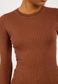 Brūns ribots ilga piedurkne tops ar apaļu kakla izgriezumu, ar teksturētu audumu un pieguļošu dizainu, kas izceļ ķermeņa siluetu.