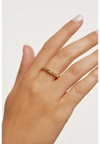 Bague en or avec des accents de pierres claires rectangulaires et rondes, montée sur une fine bande texturée, portée par une main à la peau claire.