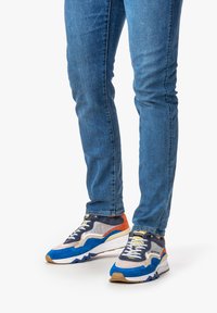 Sneaker multi-colore realizzato in mesh, camoscio e pelle. Presenta una suola prominente, un sistema di allacciatura e accenti a contrasto in blu, arancione e beige.