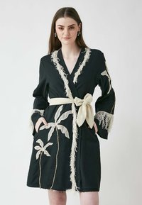 Svart kappa med fransar i beige, prydd med spetsliknande broderade palmblad. Inkluderar ett bälte och fickor, med en skräddarsydd design.