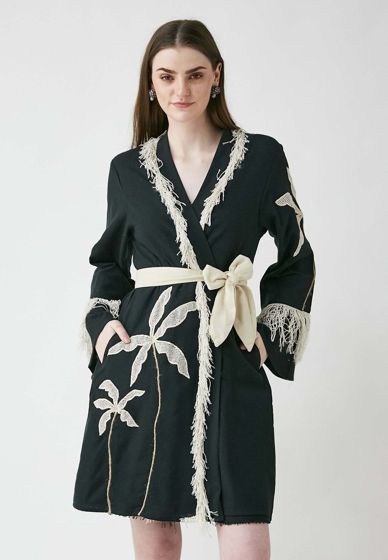 Svart kappa med fransar i beige, prydd med spetsliknande broderade palmblad. Inkluderar ett bälte och fickor, med en skräddarsydd design.