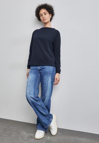 Mörkblå långärmad bomullströja, avslappnad passform, i kombination med ljusblå vida jeans och vita sneakers på en grå golv mot en neutral bakgrund.