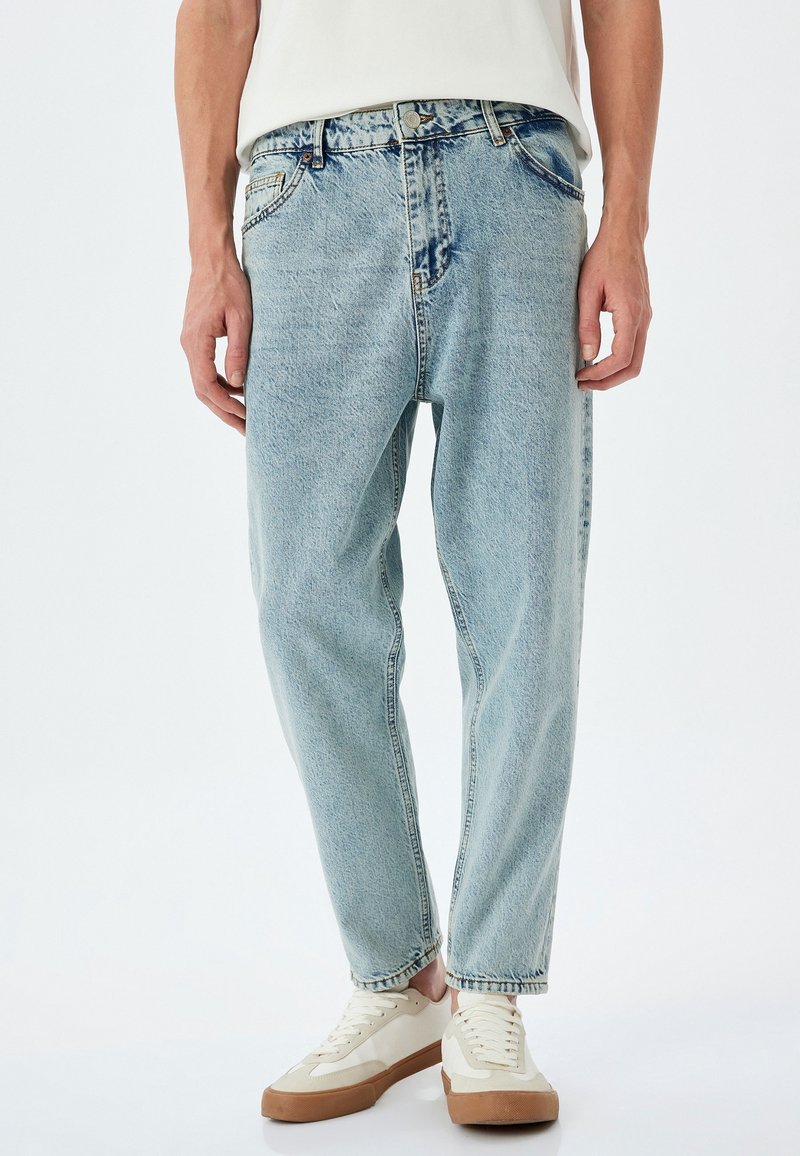 Jeans bleu clair coupe décontractée portés avec des baskets blanches et un haut blanc, visibles de la taille aux chaussures sur un fond uni.
