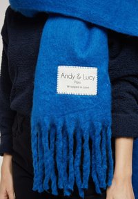 Écharpe en laine bleue avec franges, dotée d'une étiquette blanche qui indique "Andy & Lucy, Enveloppé d'Amour." Texture douce et couleur vive.