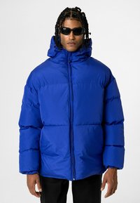 Blauwe pufferjack met een hoge kraag, ritssluiting en twee zijzakken. Beschikt over een dikke, gewatteerde structuur en elastische manchetten.
