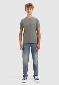 Tienerjongen staat rechtop, gekleed in een grijs T-shirt met de tekst "BALLIN'", blauwe vervaagde jeans en blauw-witte sneakers, tegen een effen lichte achtergrond.