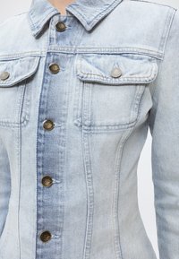 Φωτεινό μπλε τζιν μπουφάν με μεταλλικά κουμπιά και δύο τσέπες στο στήθος, εμφανίζεται από το γιακά μέχρι τη μέση σε ένα άτομο.