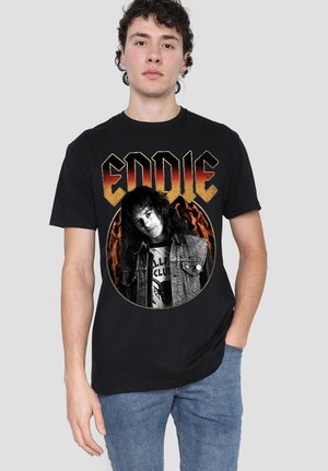 Young man wearing black "Eddie" graphic t-shirt with a black-and-white portrait inside a circle of flames, paired with blue jeans.