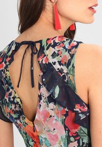 Kvinna klädd i en blommig klänning med volangärmar och ett nyckelhål på ryggen knutet med en marinblå rosett, visad från baksidan och sidan.