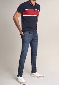 Camisa polo marina con rayas horizontales rojas y blancas, combinada con jeans de mezclilla oscura y zapatillas blancas con detalles grises.
