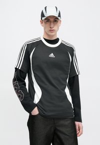 Fekete-fehér Adidas sportpóló rövid ujjakkal, amelyen a vállakon három csík látható, valamint hosszú fekete aláöltöző logómintával. A sapka a mintázathoz illeszkedik.