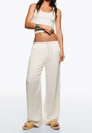 Kobieta w białym bezrękawniku typu crop top, luźnych białych spodniach z wiązaniem w pasie, srebrnej bransoletce i jasnobrązowych sandałach na płaskim obcasie z ćwiekami.