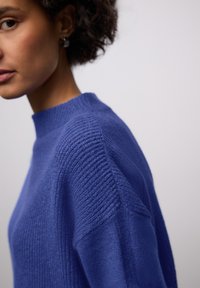 Blauer Strickpullover mit hohem Kragen, strukturiertem Rippenmuster an den Schultern und glattem Stoff am Körper. Nahaufnahme konzentriert sich auf die Nahtverarbeitung und die Form.