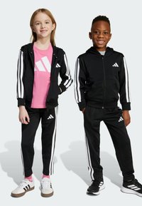 Två barn står sida vid sida iklädda svarta Adidas-träningsoveraller med vita ränder; flickan har en rosa tröja, båda ler.