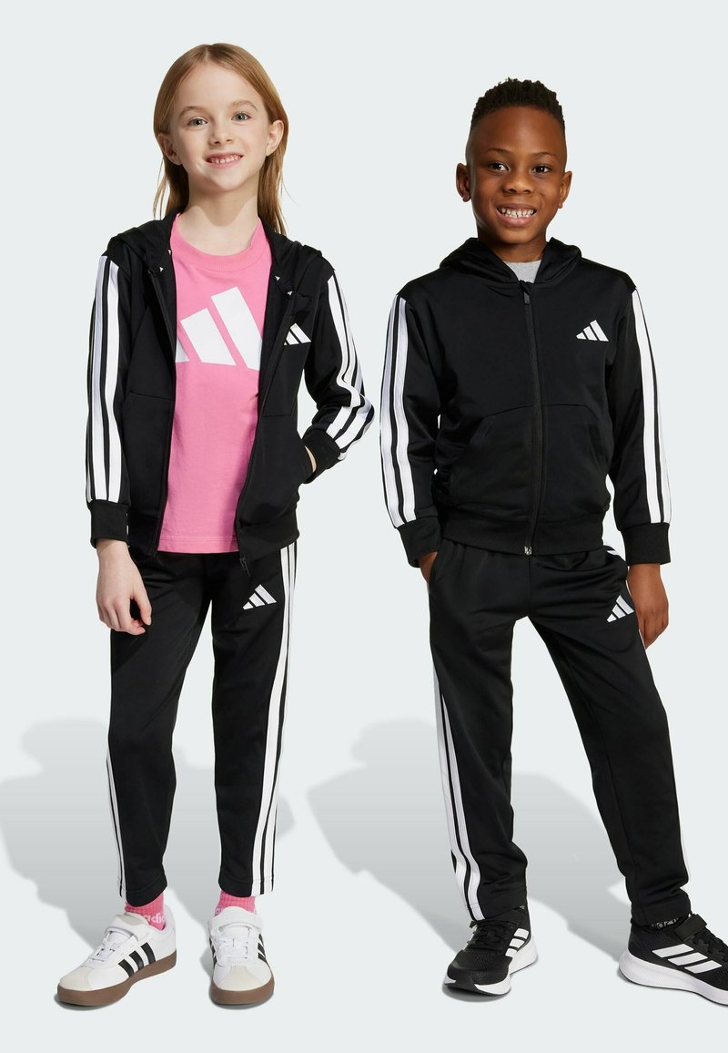 Två barn står sida vid sida iklädda svarta Adidas-träningsoveraller med vita ränder; flickan har en rosa tröja, båda ler.