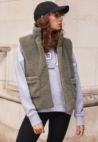 Vest fără mâneci, de culoare verde măslinie fuzzy, cu buzunare frontale, purtat peste un hanorac gri deschis, asortat cu colanți negri și o șapcă neagră.