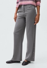 Donna che indossa pantaloni grigi a gamba larga con borchie laterali, un maglione a righe rosa e viola, e scarpe slip-on nere con borchie.