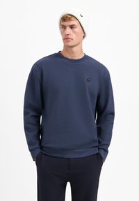 Navyblauwe sweater van gebreide stof, met een ronde hals en een klein hexagonaal logo. Gecombineerd met donkere broek en een witte gebreide muts.