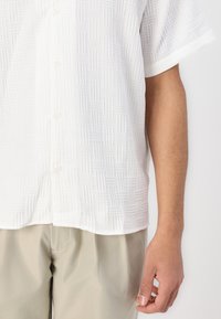 Camicetta bianca a maniche corte con un motivo geometrico testurizzato, colletto a bottoni e vestibilità rilassata, abbinata a pantaloni beige chiaro.